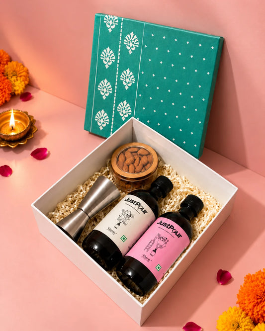 JustPour Diwali Gift Box open on a pink table with a teal lid, two bottles, a steel jigger, almonds, a lit diya and marigolds